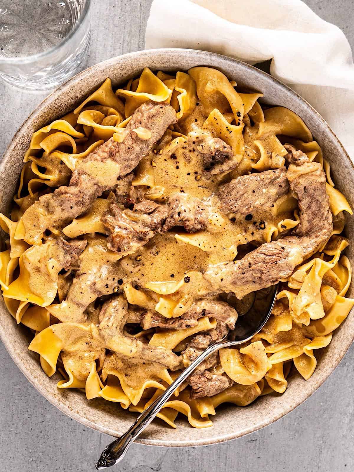 A bowl of beef stroganoff with a spoon inside and a linen napkin nearby.