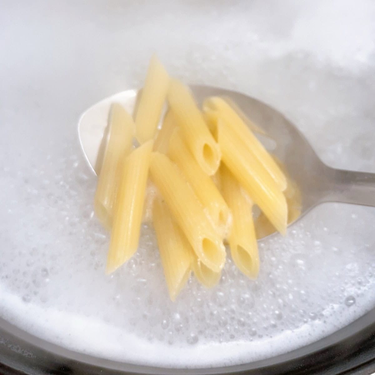 A spoon holding ziti over boiling water.