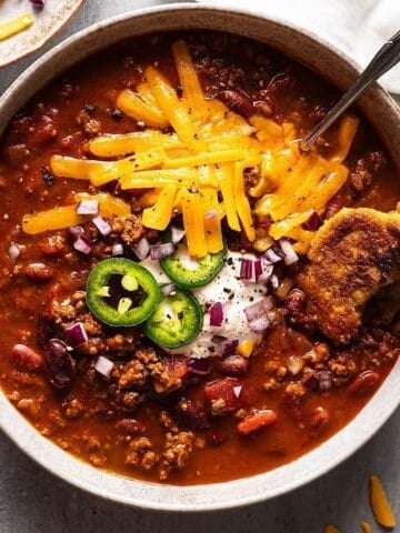 Dutch oven chili in a bowl with cheese, jalapeno, sour cream, and onion topping.