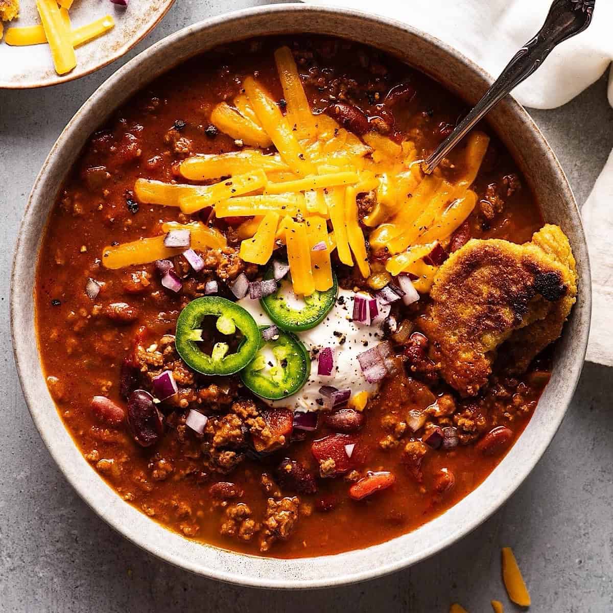 Dutch oven chili in a bowl with cheese, jalapeno, sour cream, and onion topping.