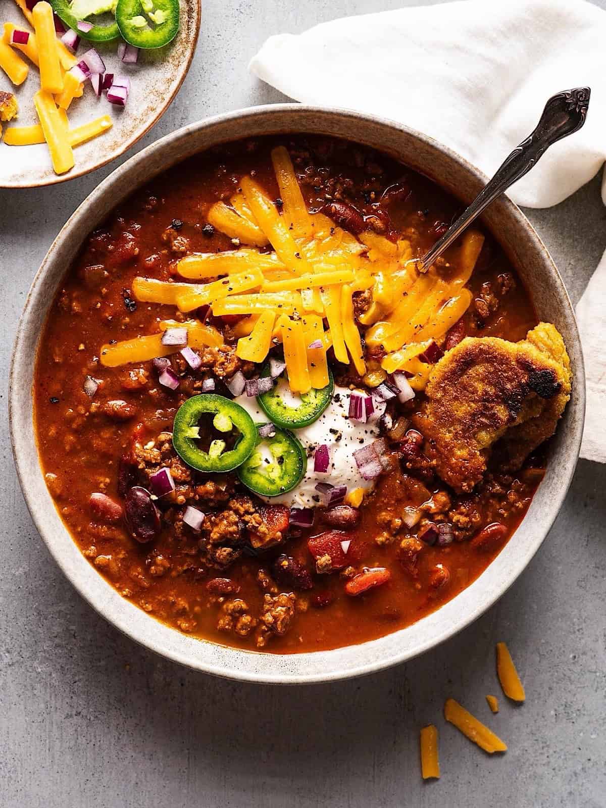 Dutch oven chili in a bowl with cheese, jalapeno, sour cream, and onion topping.