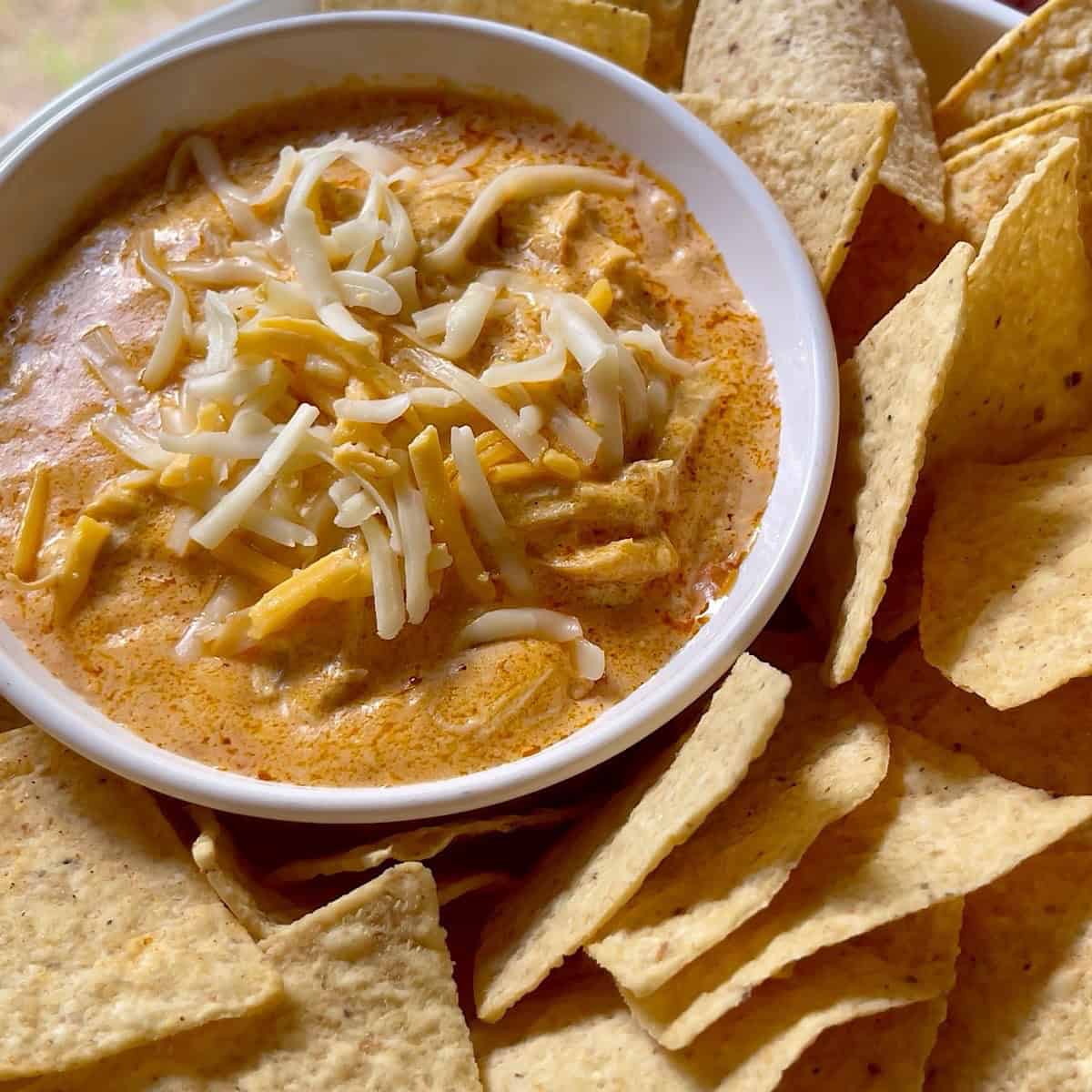 Prepared buffalo chicken dip in bowl surrounded by tortilla chips and topped with shredded cheese.