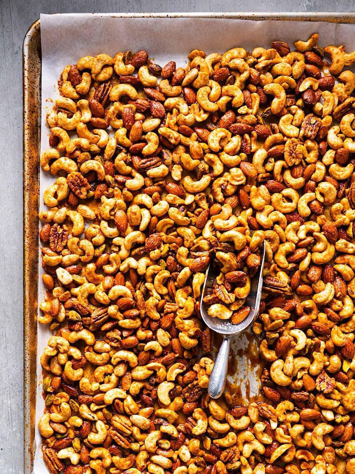 Sweet and spicy mixed nuts on a sheet pan with parchment paper and a scooper.