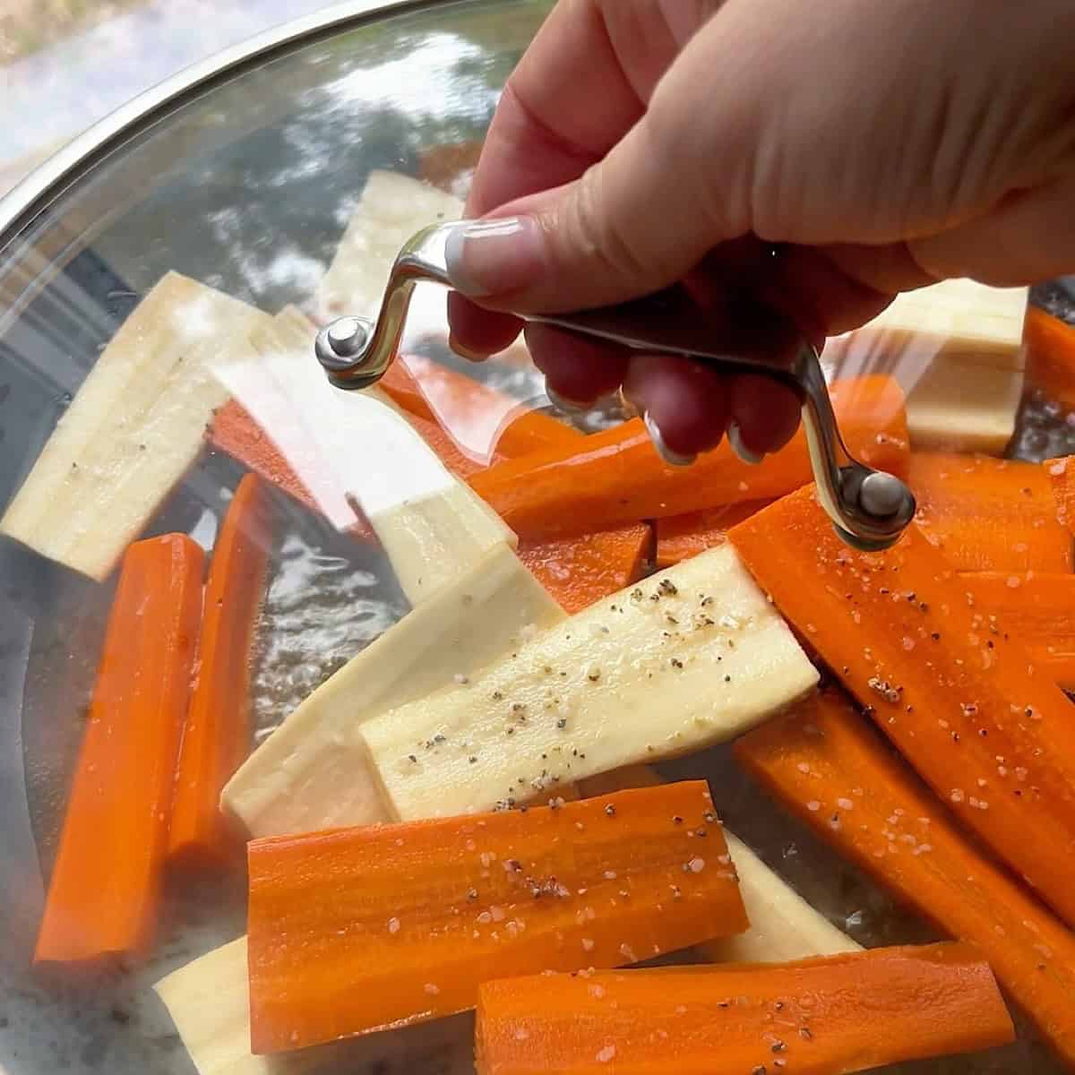 Carrot and parsnip batons simmering in broth with a glass lid on the skillet.