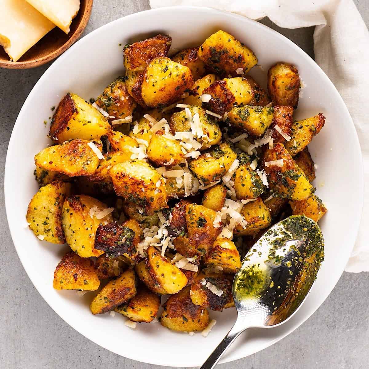 Pesto potatoes in a white bowl with a spoon, linen, and small bowl of parmesan.