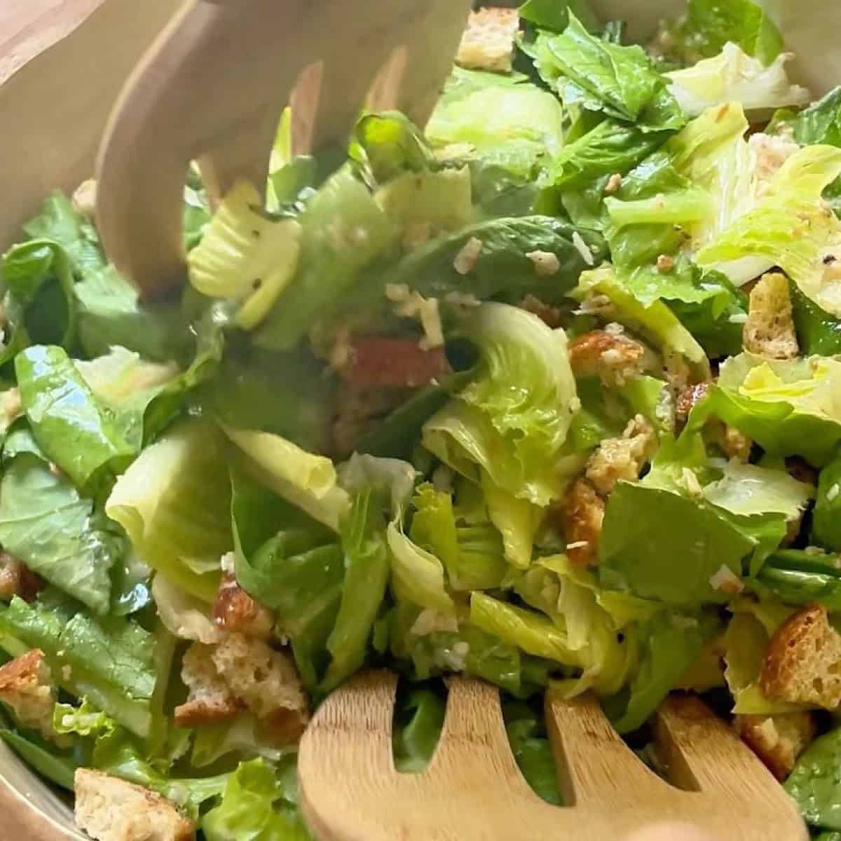 Salad being tossed with Caesar dressing, croutons, and parmesan cheese.