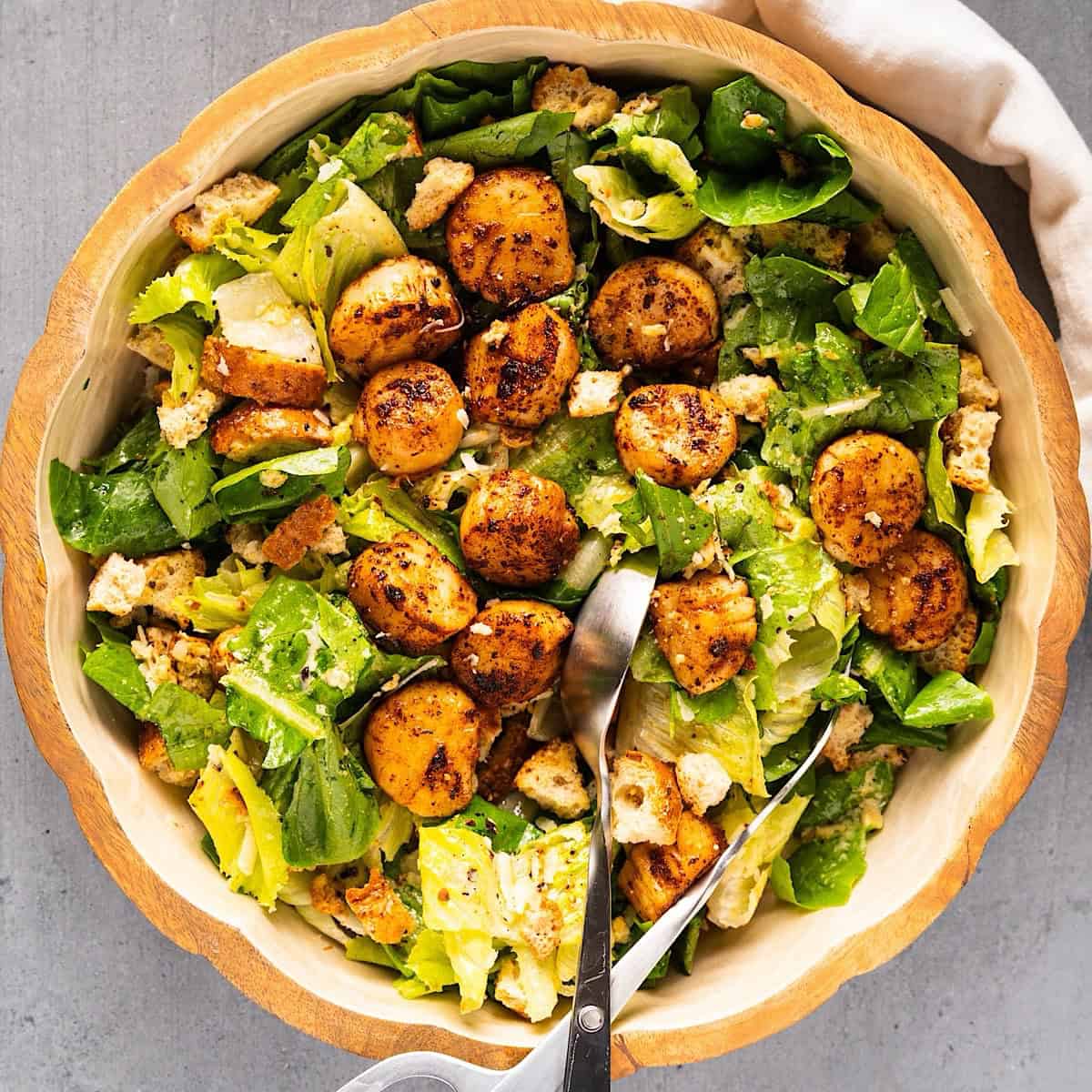 Tossed callop salad in a large salad bowl with salad tongs and linen on the side.