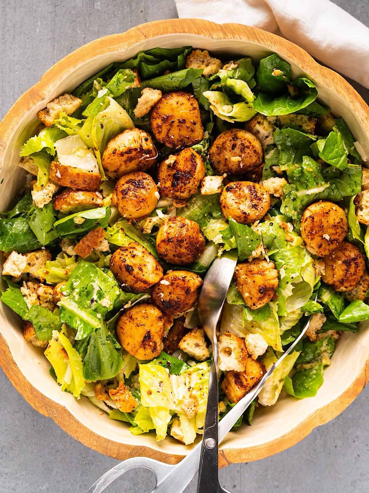 Tossed callop salad in a large salad bowl with salad tongs and linen on the side.