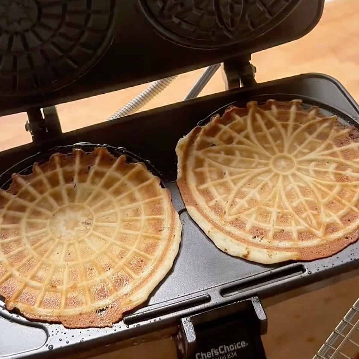 Two browned pizzelles in pizzelle maker.