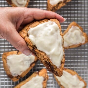 Holding a cinnamon roll scone with cream cheese glaze over a wire rack with remaining scones.