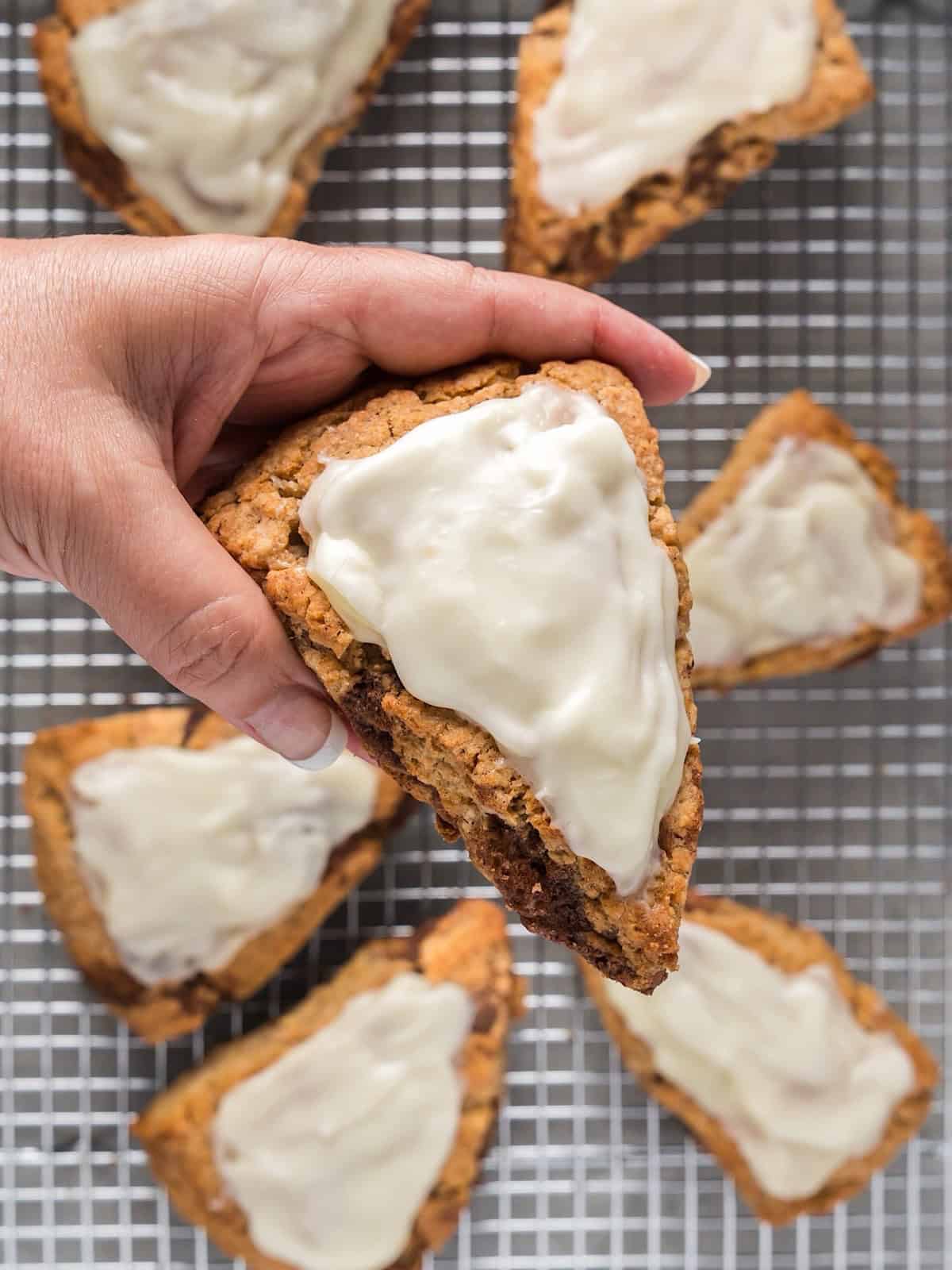 Holding a cinnamon roll scone with cream cheese glaze over a wire rack with remaining scones.