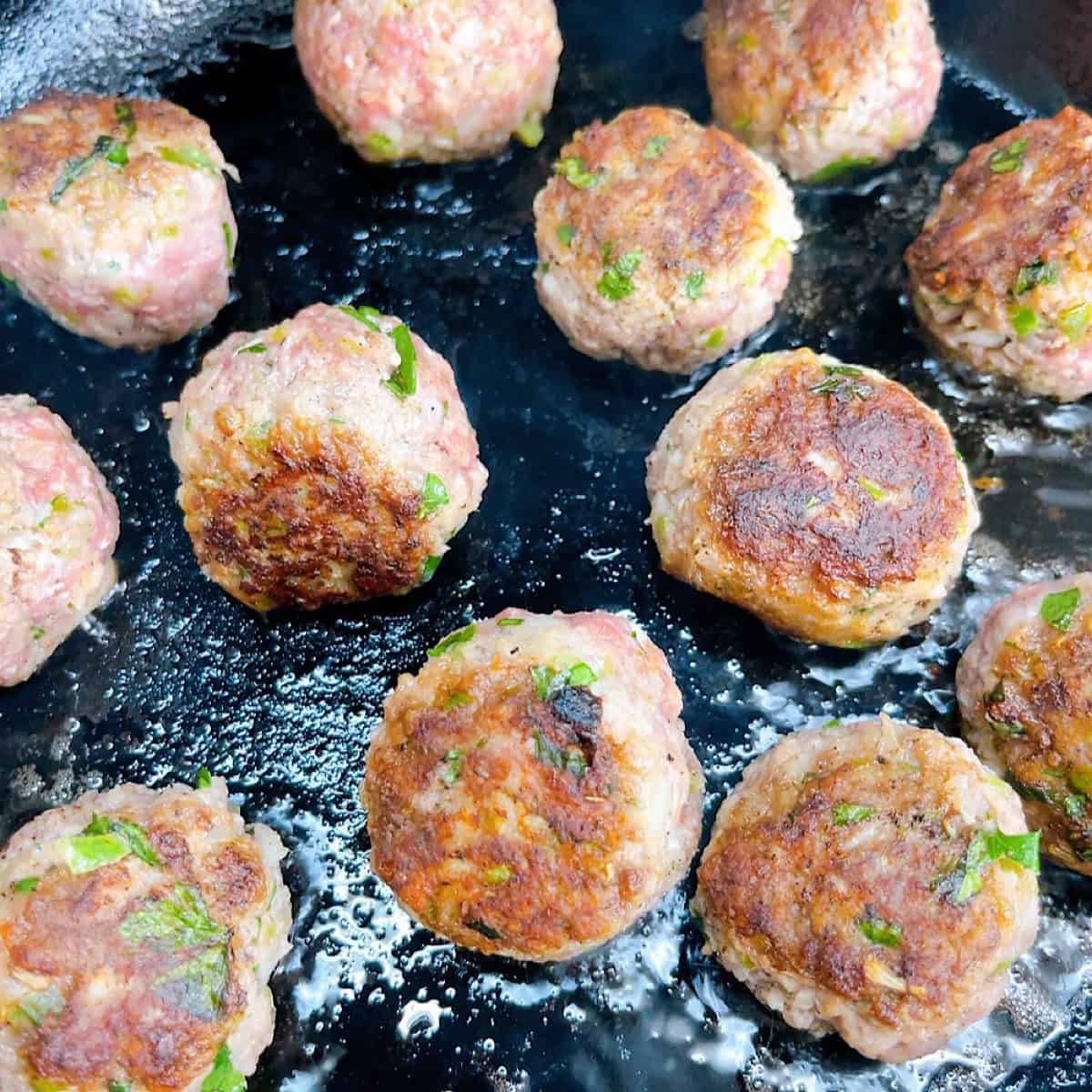 Greek lamb meatballs searing in a cast iron skillet.