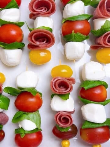 Mini caprese skewers with salami roses lined up on a platter.