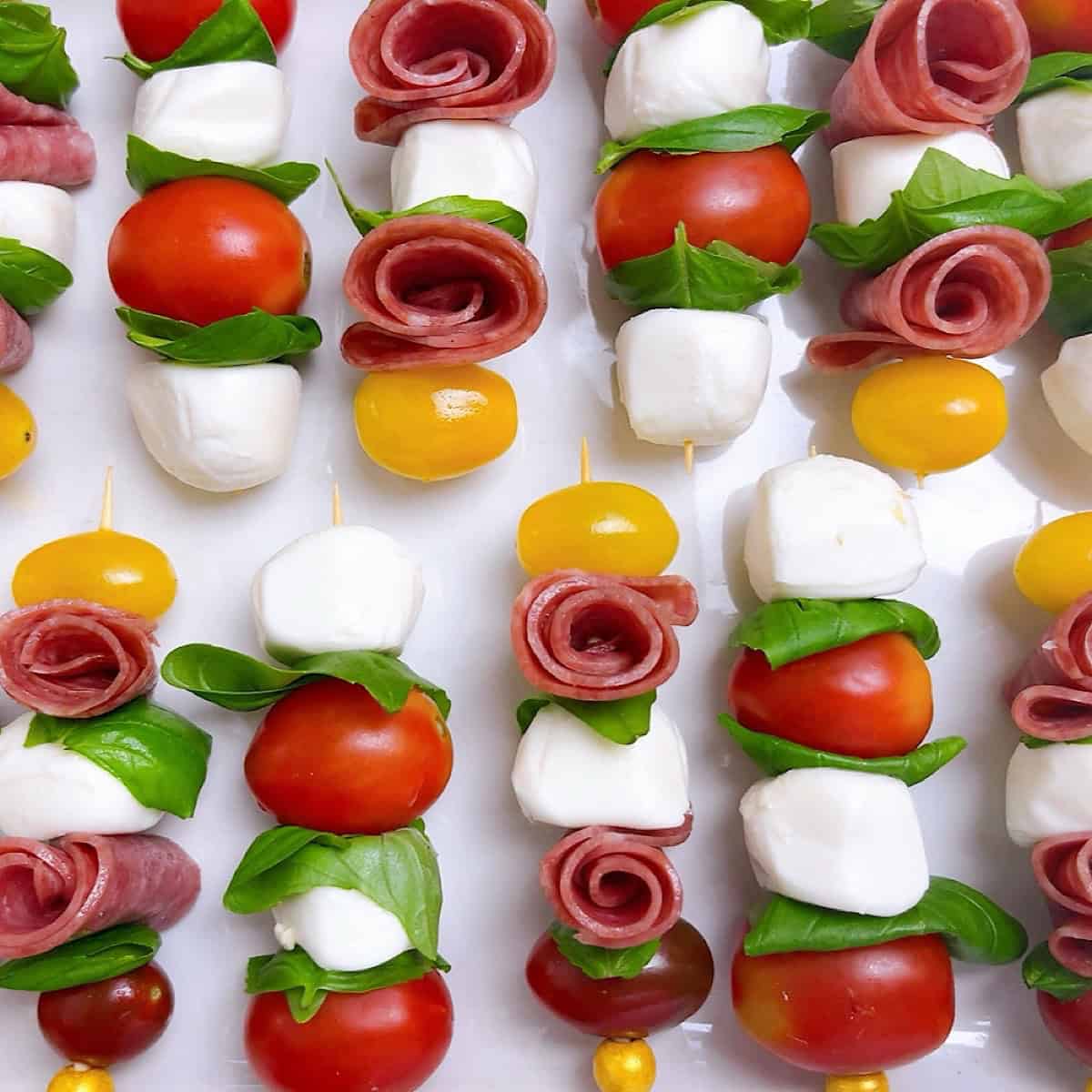 Mini caprese skewers with salami roses lined up on a platter.