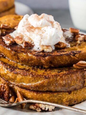 French toast with syrup and toppings stacked on a plate with milk and another stack in background.