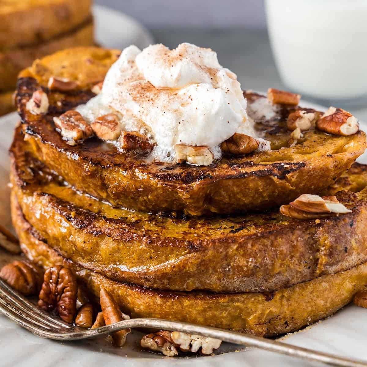 French toast with syrup and toppings stacked on a plate with milk and another stack in background.
