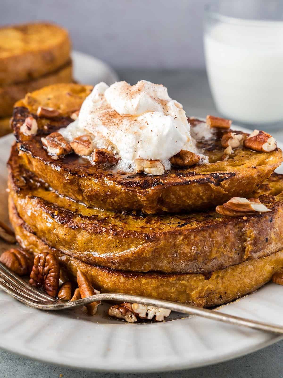 French toast with syrup and toppings stacked on a plate with milk and another stack in background.