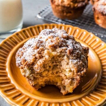 A bite shot of an apple streusel muffin with more more muffins and milk in background.