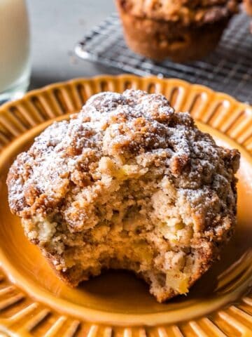 A bite shot of an apple streusel muffin with more more muffins and milk in background.