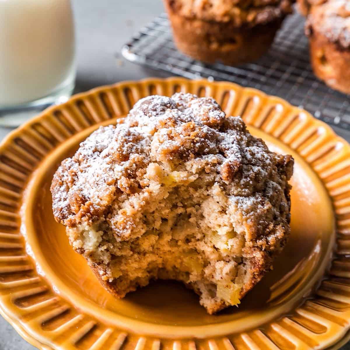 A bite shot of an apple streusel muffin with more more muffins and milk in background.