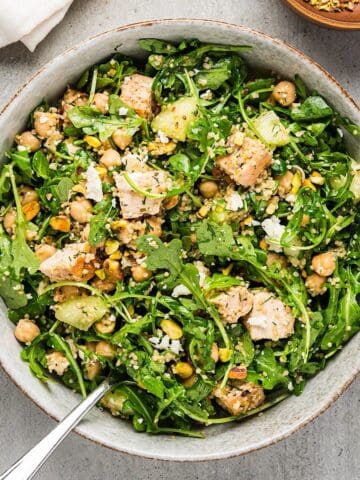 Chicken arugula salad in a wide bowl next to linen and bowl of pistachios.