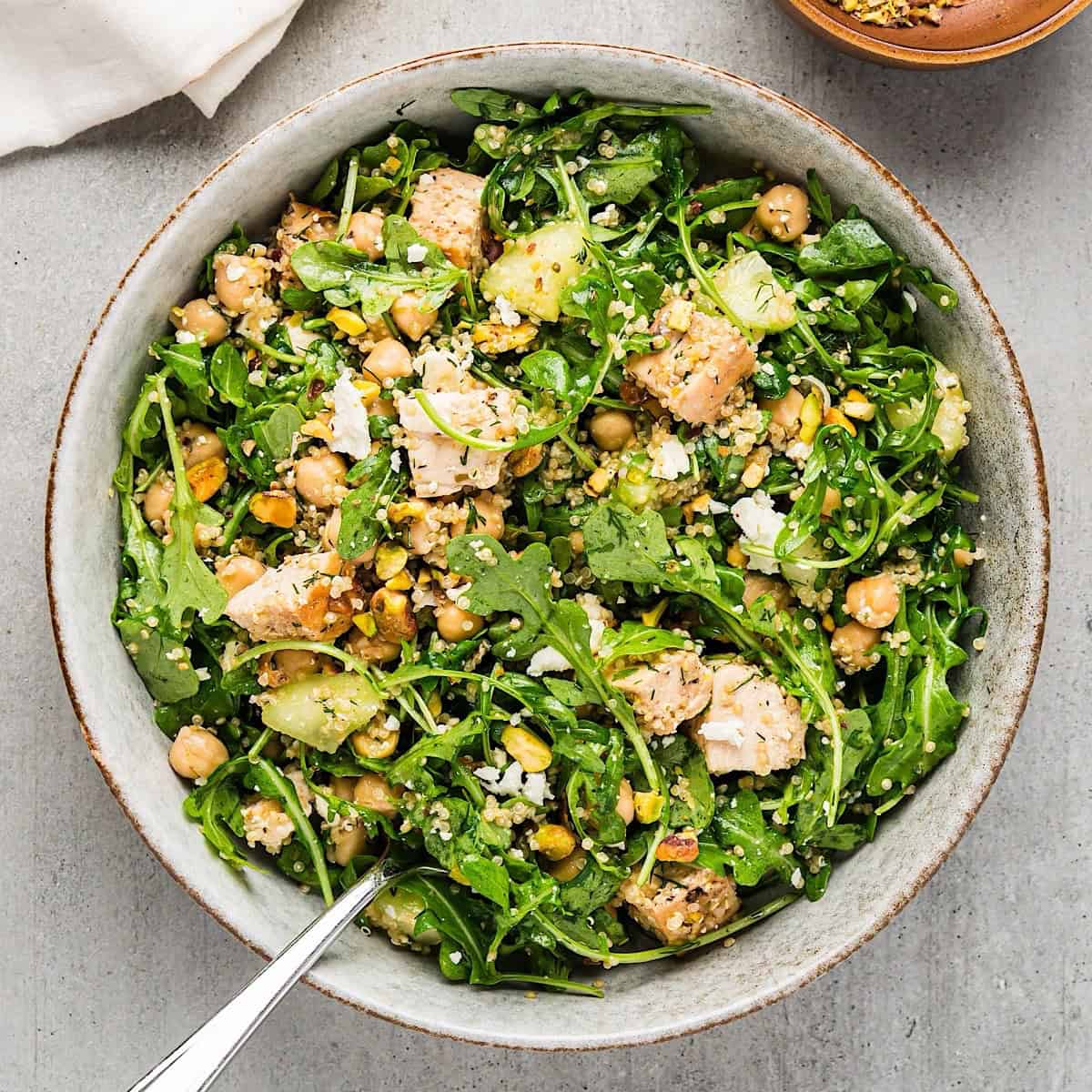 Chicken arugula salad in a wide bowl next to linen and bowl of pistachios.