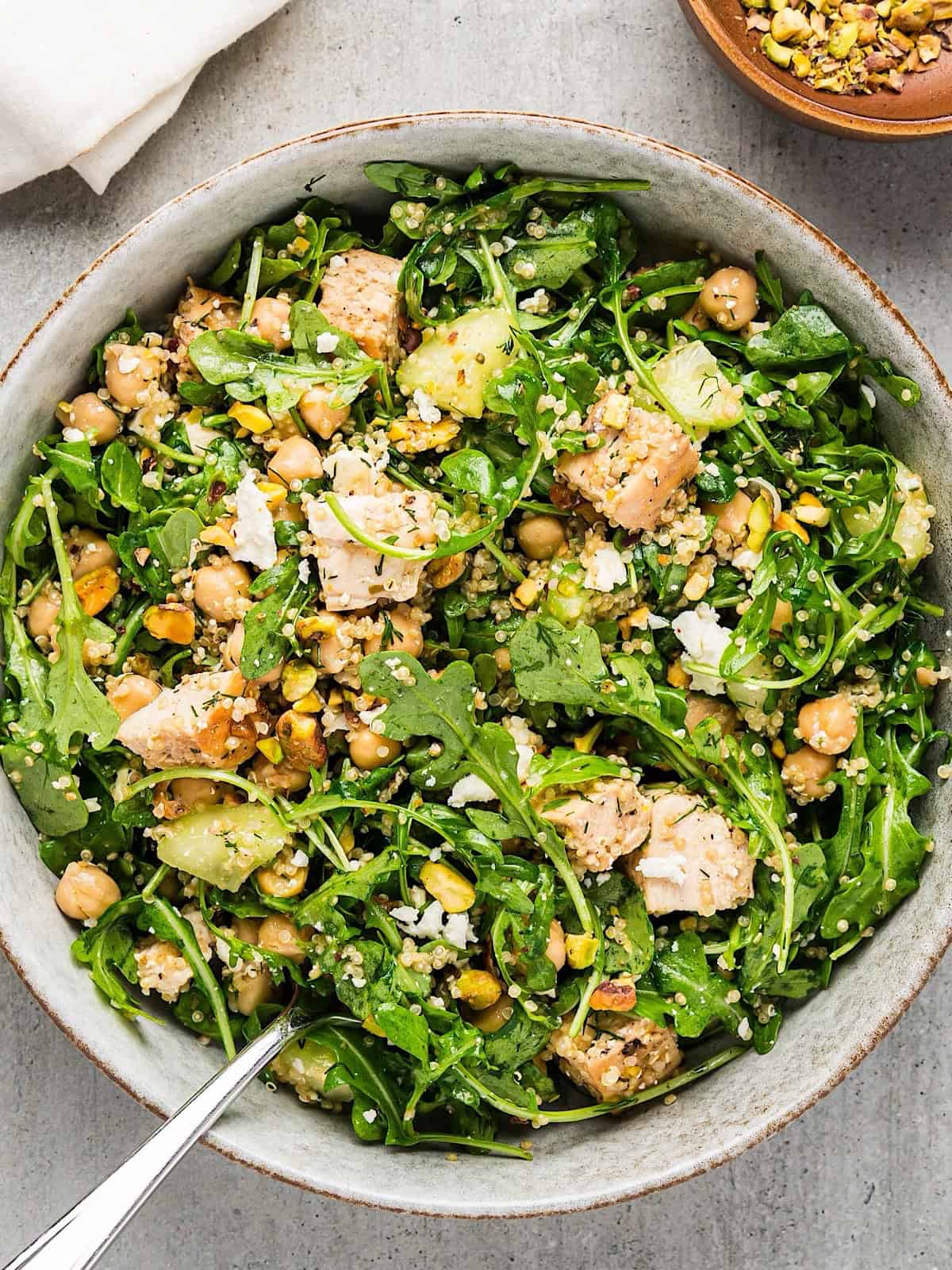 Chicken arugula salad in a wide bowl next to linen and bowl of pistachios. 