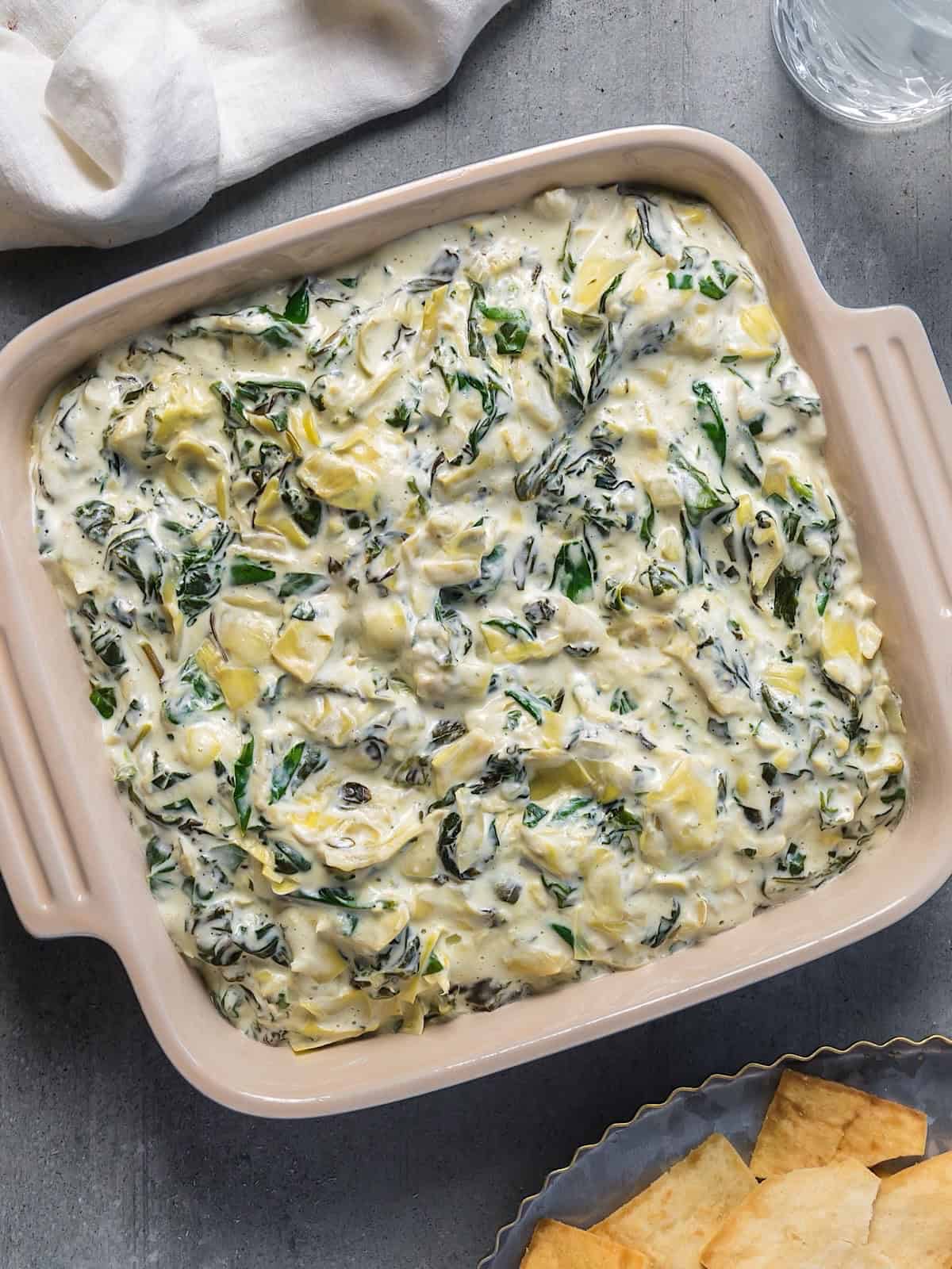 Spinach artichoke dip without mayonnaise in a ceramic dish next to a linen and glass of water.