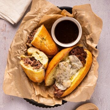 Two french dip sandwiches with a bowl of au jus gravy in a plastic basket with parchment.