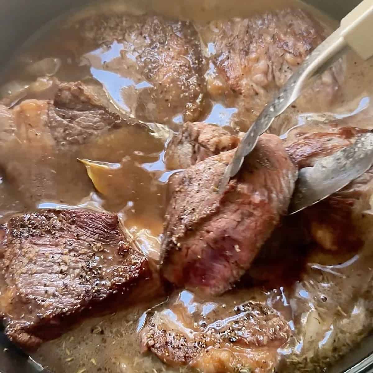 Placing the chuck roast back into the au jus beef broth base with tongs.