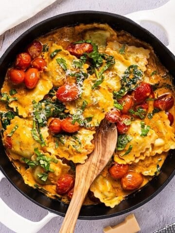Skillet ravioli with spinach and a wooden spoon near parmesan and linen.
