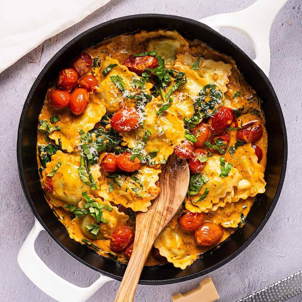 Skillet ravioli with spinach and a wooden spoon near parmesan and linen.