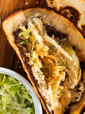 Smashed big mac tacos on wooden platter with special sauce, pickles, lettuce, and sesame seeds.