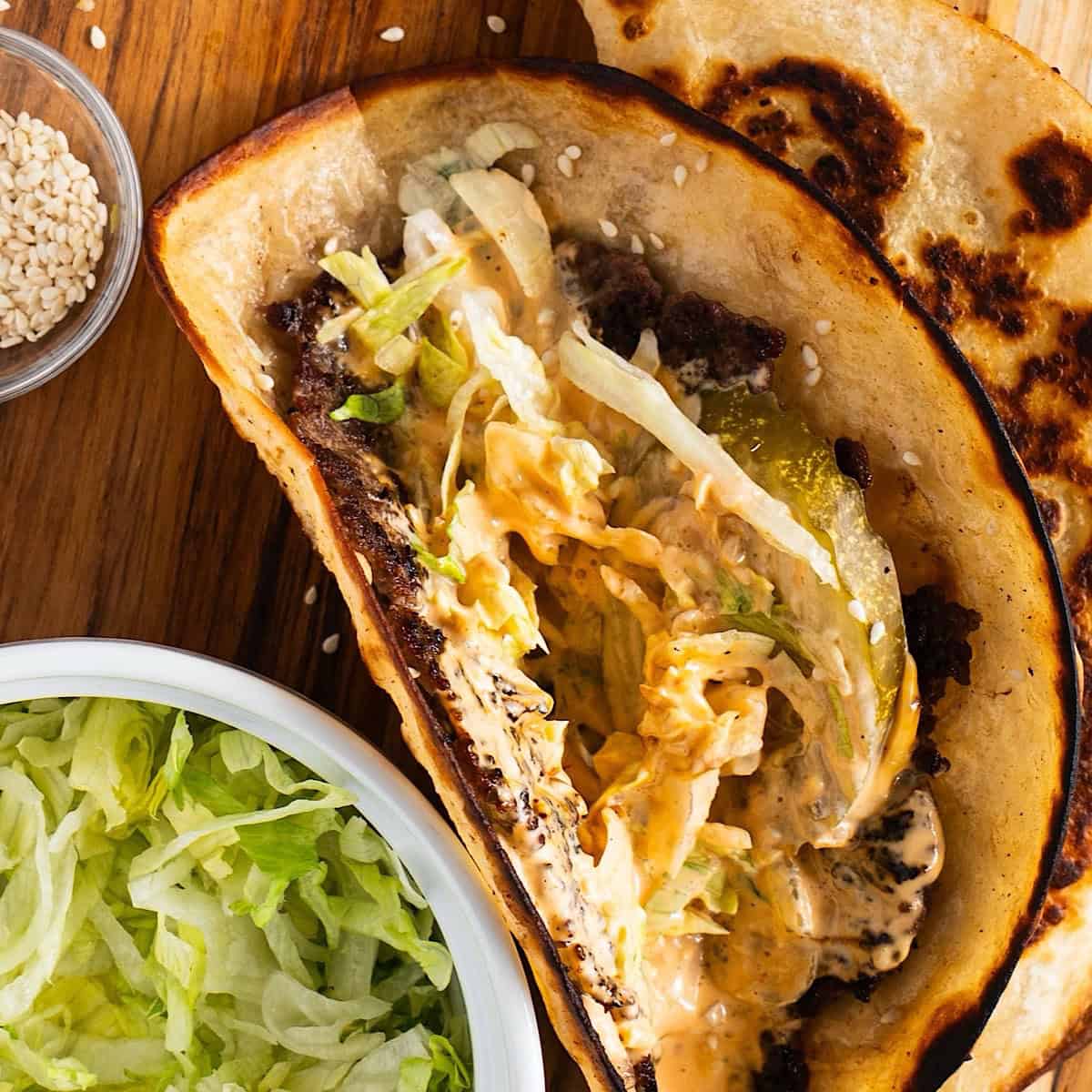Smashed big mac tacos on wooden platter with special sauce, pickles, lettuce, and sesame seeds.