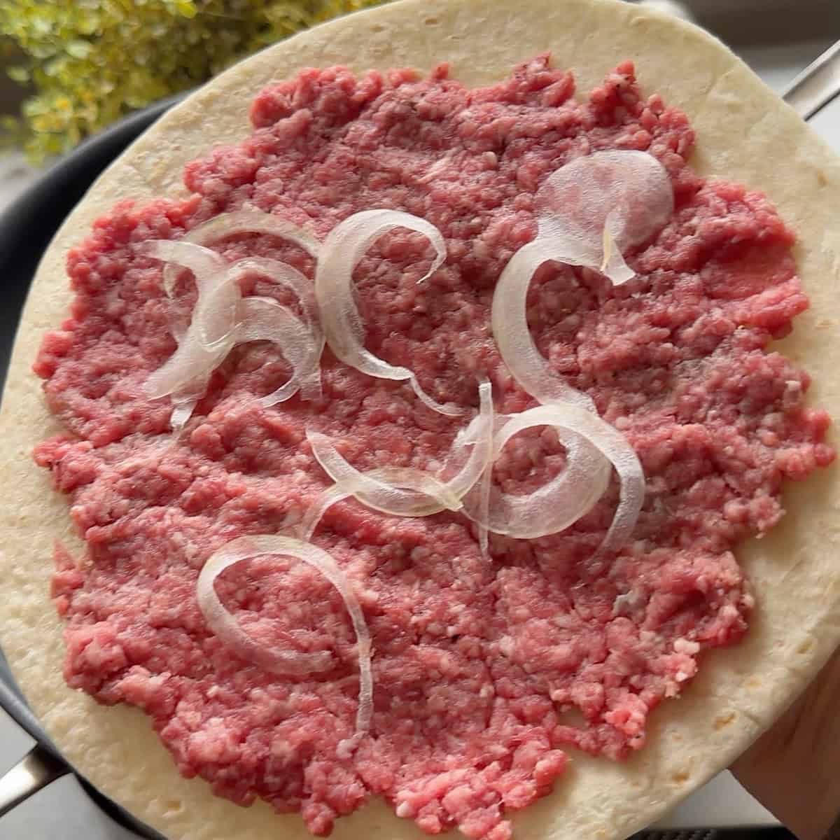 Ground beef pressed flat on a flour tortilla with onions scattered on top.