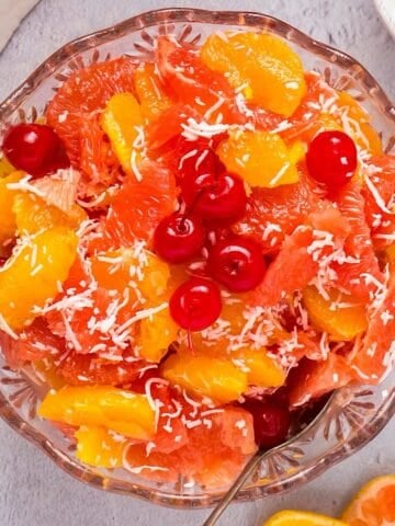 Ambrosia salad in a decorative glass bowl next to fruit, coconut, and linen.