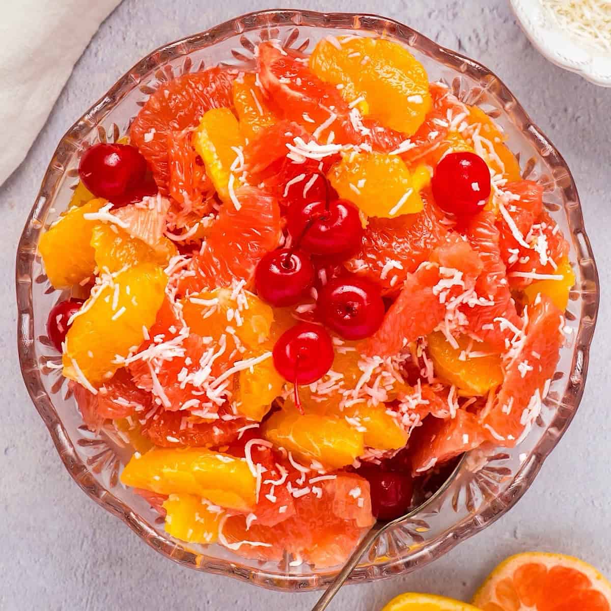 Ambrosia salad in a decorative glass bowl next to fruit, coconut, and linen.