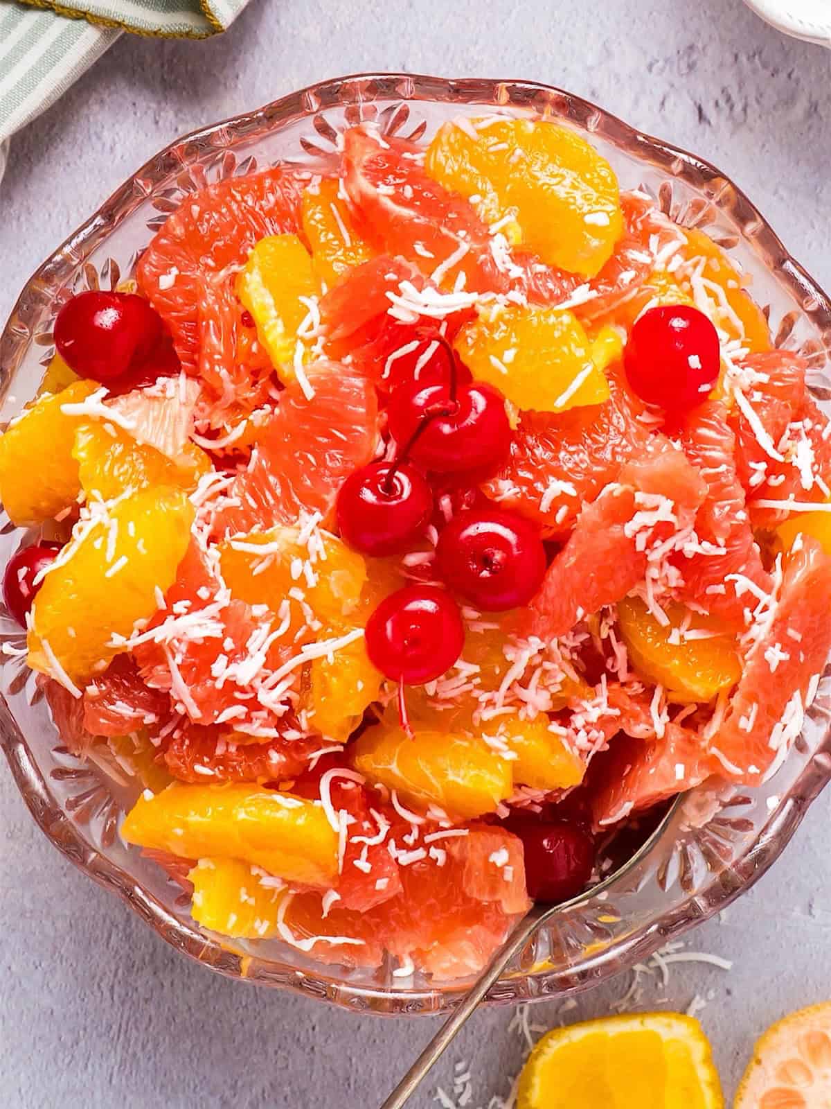 Ambrosia salad in a decorative glass bowl next to fruit, coconut, and linen.