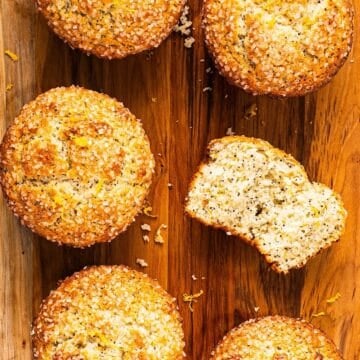 Six lemon poppy seed muffins on a wooden board with one showing the tender crumb inside.