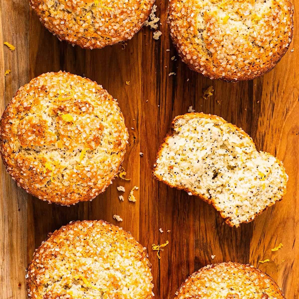 Six lemon poppy seed muffins on a wooden board with one showing the tender crumb inside.