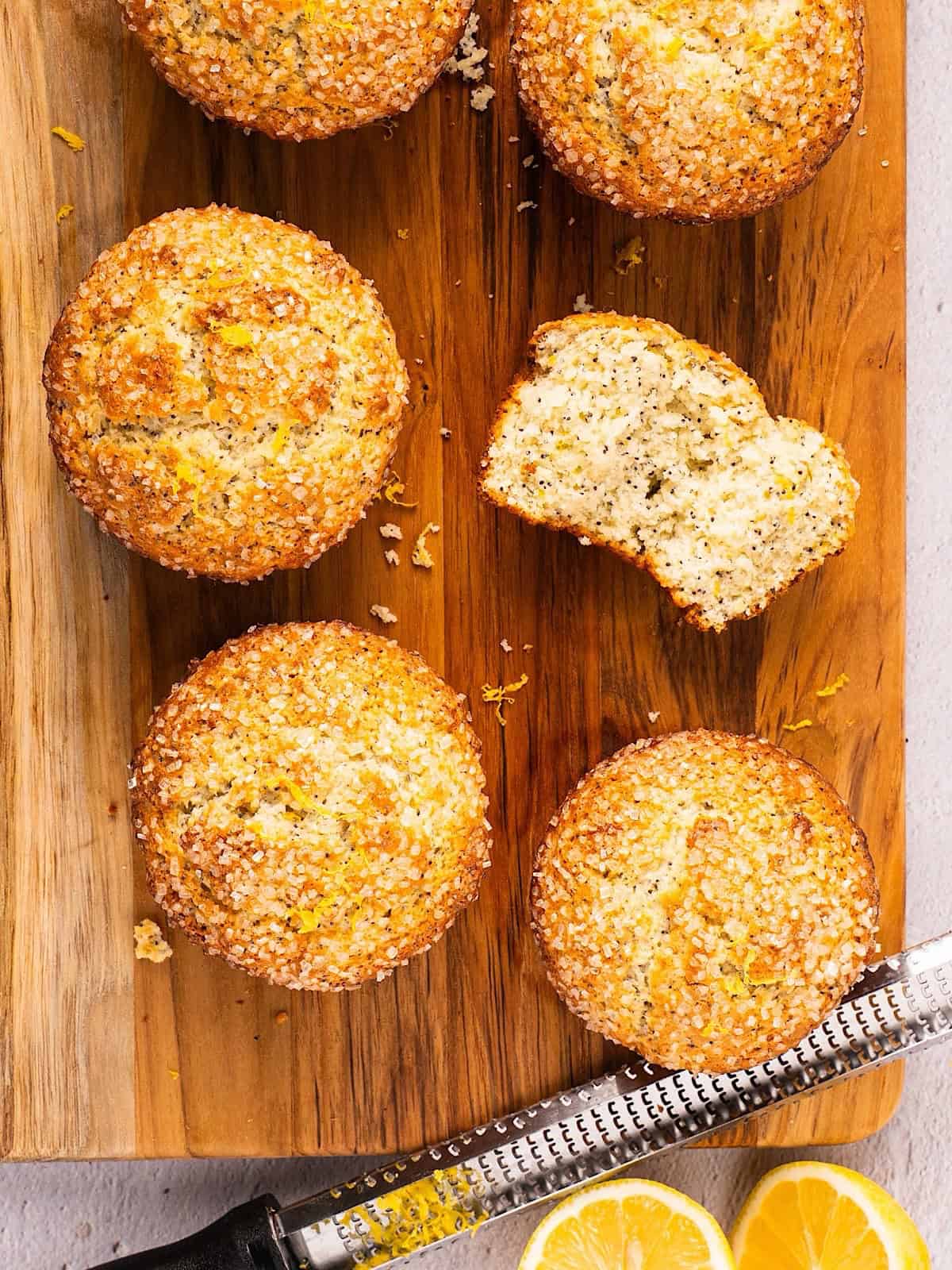 Six lemon poppy seed muffins on a wooden board with one showing the tender crumb inside.
