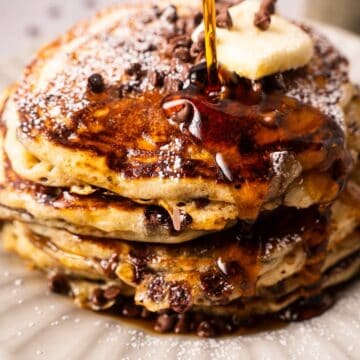 A fluffy chocolate chip pancake stack with powdered sugar and syrup cascading down the front.