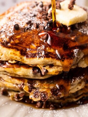A fluffy chocolate chip pancake stack with powdered sugar and syrup cascading down the front.
