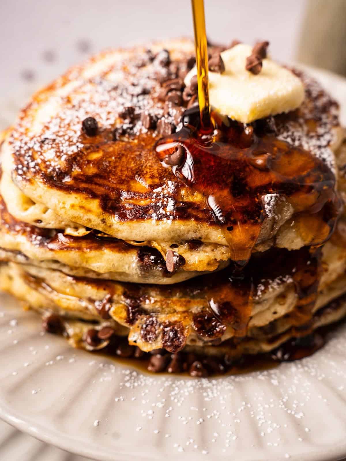 A fluffy chocolate chip pancake stack with powdered sugar and syrup cascading down the front.