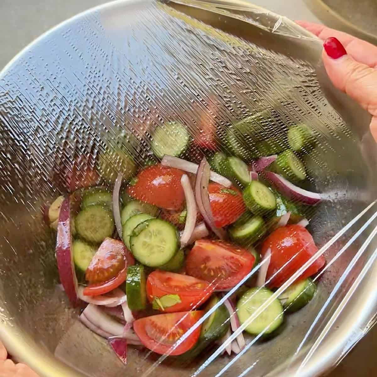 Covering the tomato cucumber salad with plastic wrap to marinate and chill.