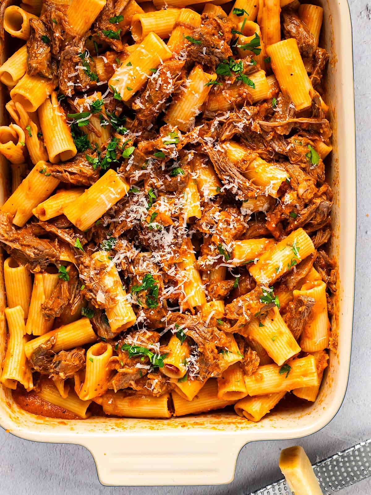 Beef ragu rigatoni in a casserole dish with parmesan and parsley garnish.