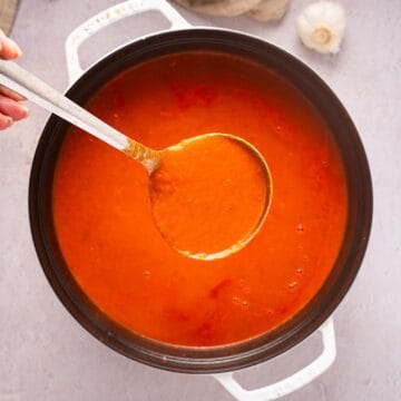 Holding a ladle of marinara sauce over a white dutch oven with garlic and linen cloth in background.