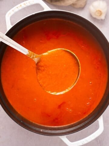 Holding a ladle of marinara sauce over a white dutch oven with garlic and linen cloth in background.