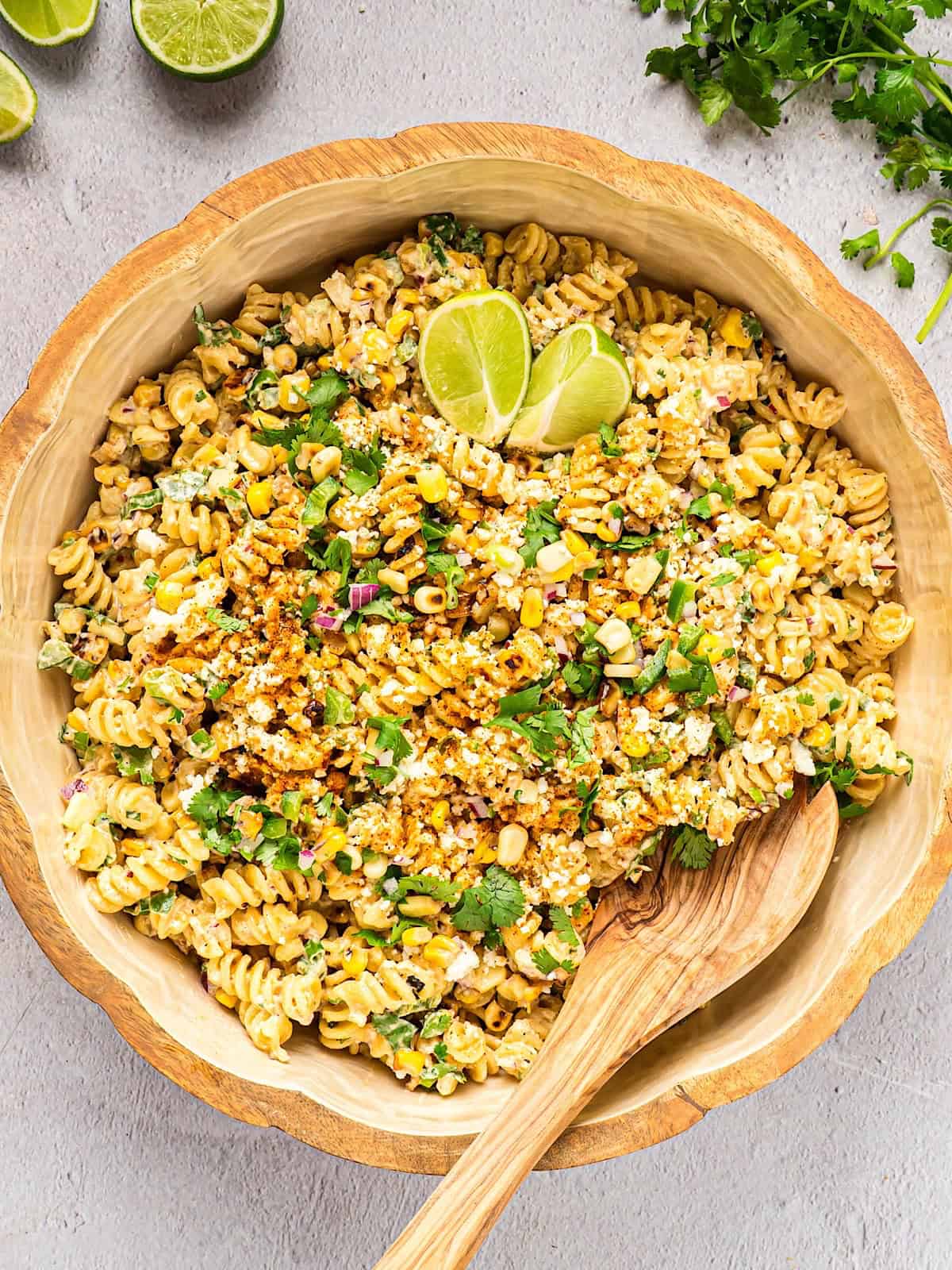 Elote pasta salad in a wooden bowl with a serving spoon and lime cilantro garnish.