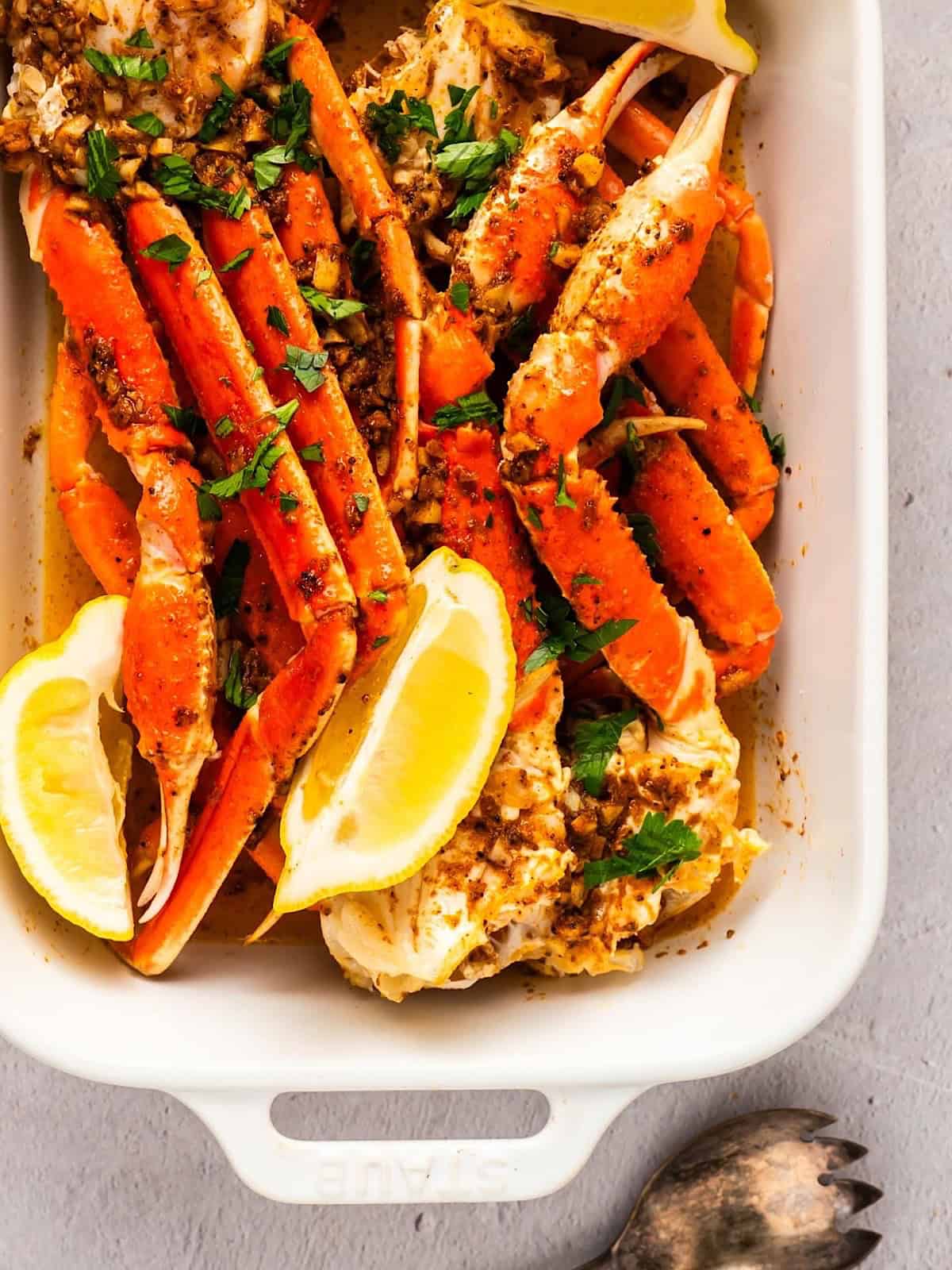 Oven baked snow crab legs in a baking dish with lemon wedges, parsley, and garlic butter.